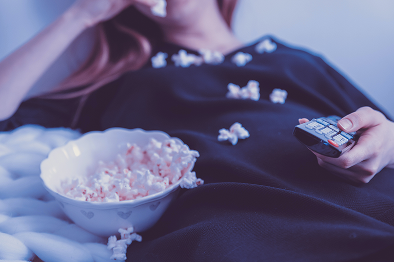How Eating Late at Night Could Be More Harmful Than You Know, image of a woman eating popcorn on the couch with a tv remote